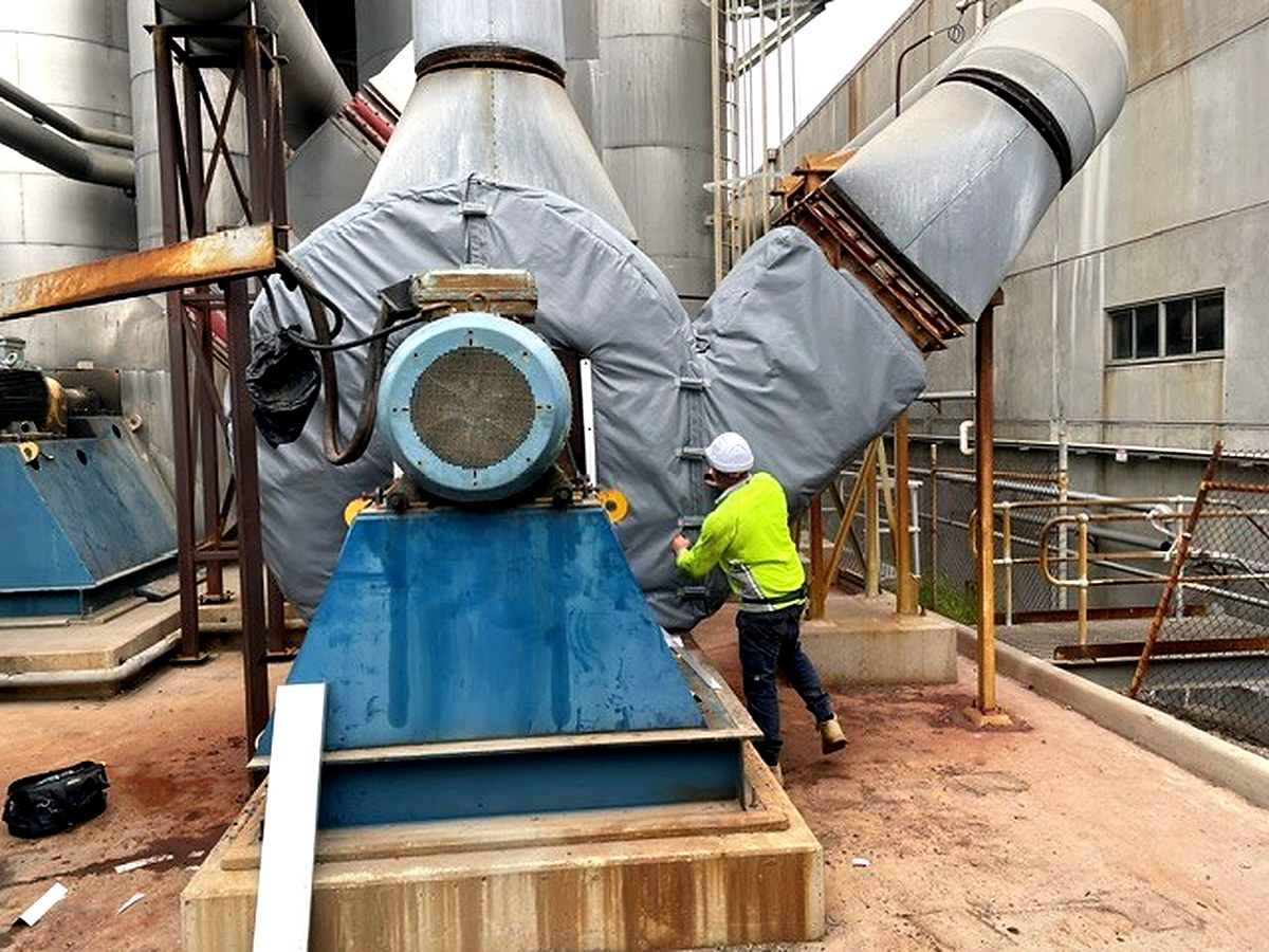 Worker fitting a large thermal jacket to an industrial fan housing at an outdoor industrial site